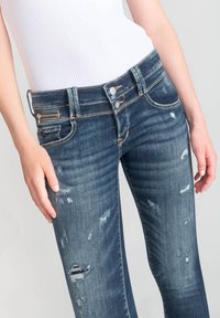 Jeans de mezclilla azul oscuro con un ajuste slim, ligero desgaste y un bolsillo con cremallera en la parte frontal. Tejido de algodón con detalles de costura en naranja.