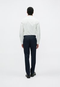 Fehér hosszú ujjú ing gombos mandzsettával és sötétkék nadrág, két hátsó zsebbel és sima, lapos textúrával. Fekete cipővel viselve.