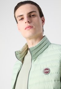 Chaqueta acolchada verde claro con costuras horizontales, cuello alto y cierre con cremallera. Presenta un parche redondo de logo marrón en el pecho.
