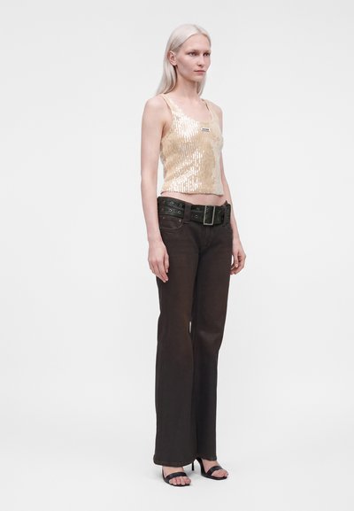 Femme aux longs cheveux blonds portant un débardeur beige à sequins, un jean large et foncé avec une grande ceinture, ainsi que des sandales noires à talons sur fond blanc.