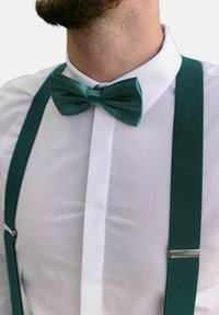 Teal sammetfluga på en vit skjorta, i kombination med mörk teal hängslen, som har en texturerad yta och silverfärgade metalldetaljer.