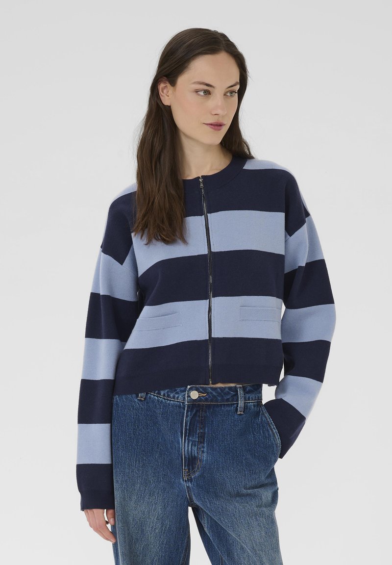 Maglione a zip a righe blu e blu navy con scollo rotondo, dotato di due tasche frontali e un design cropped. Indossato con jeans di denim a vita alta.