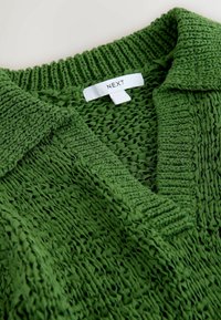 Pull vert en tricot avec col côtelé et étiquette blanche portant l'inscription « NEXT » au niveau de l'encolure.