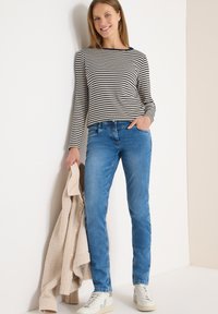 Haut à manches longues à rayures crème et marine, jean en denim bleu, tenant un cardigan en maille beige, et portant des baskets blanches avec des accents verts.