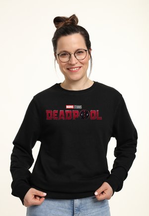 Kvinde med briller og håret i en knold smiler, mens hun har en sort Marvel Deadpool sweatshirt og lyseblå jeans på mod en hvid baggrund.