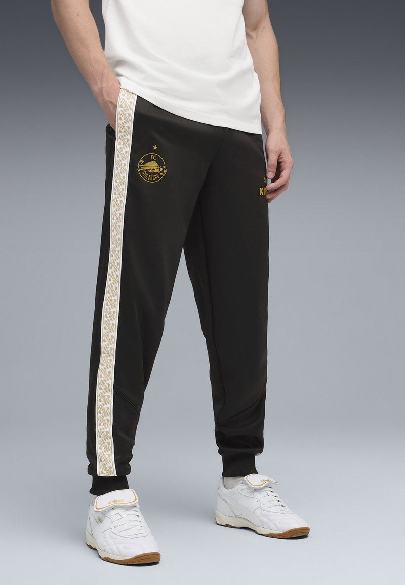 Zwarte sportbroek met een wit en goud patroon aan de zijkant. Bevat het FC Salzburg-logo in goud. De broek heeft elastische boorden en zakken.