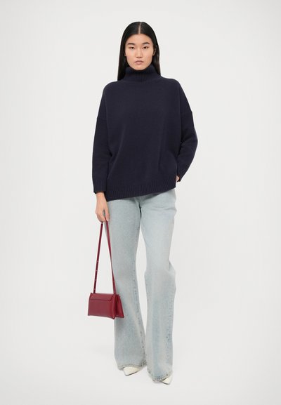 Navy turtleneck sweater, ribbed cuffs and hem, paired with light blue flared jeans and a small burgundy handbag, standing on a plain background.