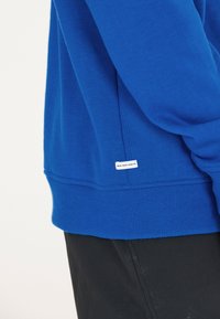 Blauwe sweatshirt met een geribbelde zoom en boorden, voorzien van een klein label met "NEW BAD HABITS" aan de zijkant. Gemaakt van een zachte stof.