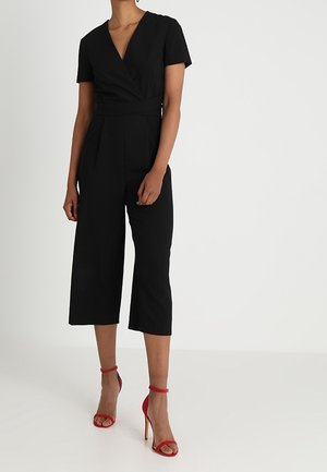 Zwarte jumpsuit met een diepe V-hals, korte mouwen en een wikkelontwerp. Culotte-stijl broekspijpen die halverwege de kuit eindigen. Gecombineerd met rode stilettohakken.