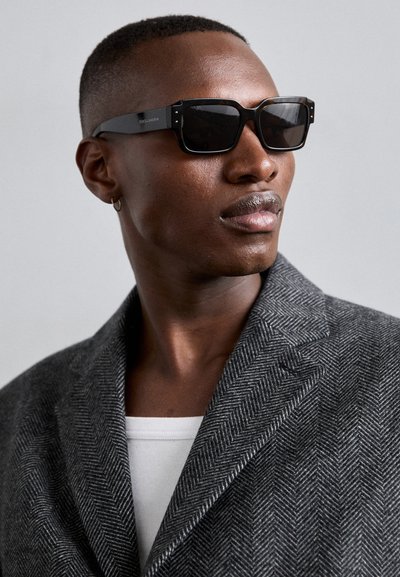 Gafas de sol negras con montura gruesa y brillante. Se lleva un blazer gris con patrón de espiga sobre una camisa blanca. Textura de piel suave visible.