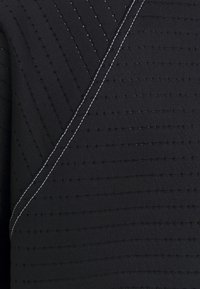 Haut en tissu noir avec des rayures verticales texturées et des détails fins de couture. Un fil blanc contrastant souligne une couture diagonale.