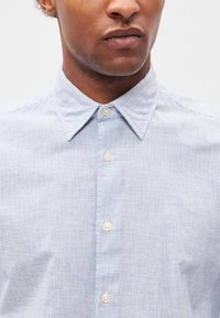 Ανοιχτό μπλε πουκάμισο με ρίγες, κουμπωτό, φτιαγμένο από ύφασμα υφαντό. Διαθέτει κλασικό γιακά και λευκά κουμπιά. Κοντινή λήψη της �περιοχής του στήθους.