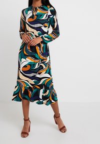 Sukienka z długim rękawem w wielokolorowy wzór z wirującymi wzorami w zielonym, pomarańczowym i kremowym, z falbaną u dołu i szczegółem z wiązanym dekoltem.