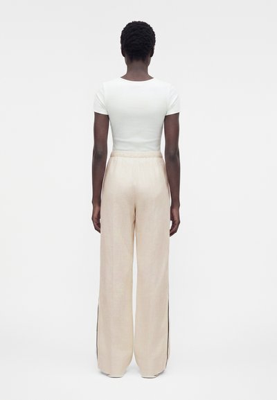 Γυναίκα που στέκεται με την πλάτη, φοράει στενό λευκό κοντομάνικο τοπ και φαρδιά μπεζ παντελόνια με ελαστική μέση.