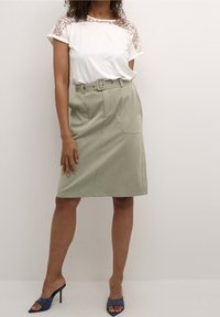 Valkoinen t-paita, jossa on pitsiyksityiskohtia olkapäillä, yhdistettynä korkeavyötäröiseen, vaaleanvihreään vyöllä varustettuun hameeseen ja sinisiin avokkaisiin.