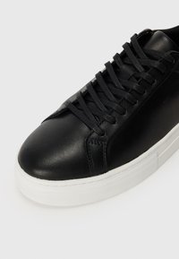 Sneaker in pelle nera con punta arrotondata, suola in gomma bianca e lacci neri piatti. Presenta una superficie liscia e un design minimalista.