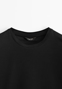 T-shirt noir à col rond avec étiquette de cou visible montrant le nom de la marque, posé à plat sur un fond blanc.
