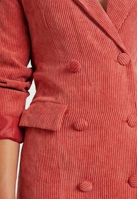Dvouřadový kordový kabát v korálově červené barvě, se žebrovanou texturou, kulatými látkovými knoflíky a ohrnutým rukávem s kontrastním akcentem.