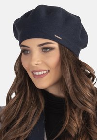 Sötétkék kötött beret, texturált felülettel és kis fém díszítéssel. Puha anyag laza esést biztosít. A modellen fekete garbó van.