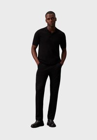 Polo negru, pantaloni negri și pantofi negri. Design slim fit, textură netedă, detalii minime, purtat cu mâinile în buzunare.