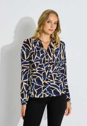 Mujer con camisa de manga larga abotonada con un patrón abstracto en azul marino, beige y negro, combinada con pantalones negros, de pie frente a un fondo claro.