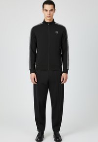 Chaqueta negra con cremallera y franjas blancas en los costados, combinada con pantalones negros de corte estrecho. Presenta un logo sencillo en el pecho y una textura elegante. Iconos en las mangas.