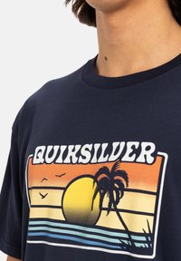 Camiseta de algodón en color navy con un estampado gráfico que incluye un atardecer, palmeras y pájaros. Cuello redondo y mangas cortas.