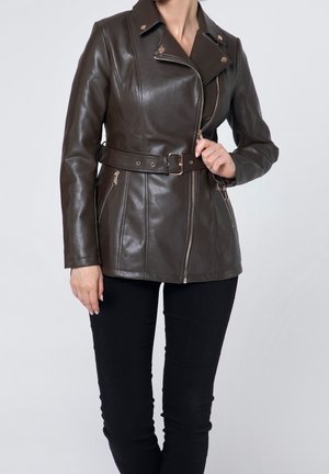 Femme portant une veste en cuir marron foncé avec ceinture, fermeture éclair asymétrique, et pantalon noir, tenant la fermeture éclair de la veste près de la poitrine.