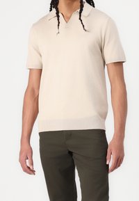 Beige kortärmad pikétröja med V-ringning, stickad textur och ribbat ärmslut. Matchas med olivgröna byxor som visar en avslappnad passform.