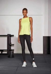 Vrouw in een limegroene mouwloze top en zwarte leggings die in een sportschool staat met gewichten en oefenmattes op de achtergrond.