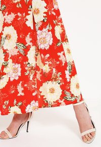 Robe longue à fond rouge vif ornée de grands motifs floraux blancs et roses, associée à une sandale à talons claire à brides.