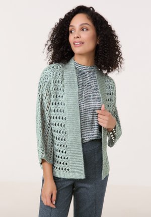 Hellgrüne Strickjacke mit Lochmuster, Schalkragen und dreiviertellangen Ärmeln. Getragen über einer gemusterten Bluse und eleganten Hosen.