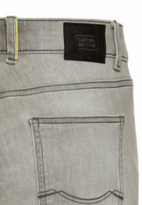 Graue Jeans aus Denim mit einem dunklen Etikett, das "camel active" zeigt, gelben Nähten und einem typischen Design der Gesäßtaschen. Glatte Textur und klassischer Schnitt.