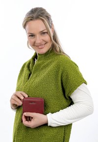 Lächelnde Frau in einem grünen, strukturierten Schal und weißem Langarmshirt hält eine kleine rote Geldbörse mit Reißverschluss vor einem weißen Hintergrund.