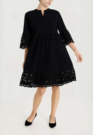 Donna che indossa un abito nero al ginocchio con rifiniture in pizzo sangallo su maniche e orlo, abbinato a scarpe nere con tacco basso, in piedi su sfondo bianco.