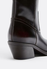 Rjava usnjena gležnjarica z rahlo ukrivljenim blok petom, detajlno šivanjem in gladko teksturo.