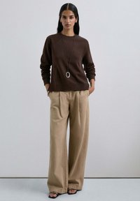 Sudadera marrón con puños acanalados, combinada con pantalones de pierna ancha beige. Un collar largo de péndulo plateado agrega un toque de detalle.
