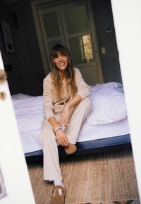 Set de haine confortabile bej, cu bluză cu fermoar și pantaloni largi. Modelul stă pe un pat cu lenjerie de pat albă, purtând papuci maro pe un covor împletit.