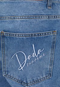 Kék farmernadrág hátsó zsebe fehér hímzett „Doda supreme” felirattal a zsebén.