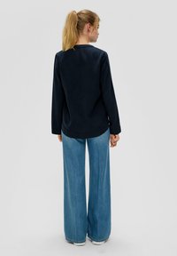 Marineblauwe blouse met lange mouwen en een ronde halslijn, gecombineerd met brede, lichtblauwe jeans met zichtbare stiksels en zoomdetails. Achteraanzicht.