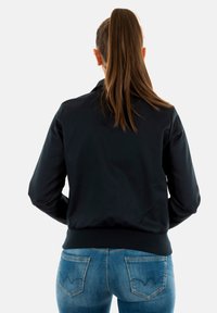 Zwarte bomberjack met een geribbelde zoom en manchetten. Glanzende stof en een kraag. Draagt een lichtblauwe jeans met een subtiele achterzakontwerp.