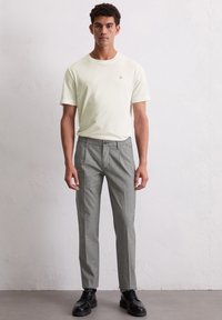 Mann, der ein weißes T-Shirt mit kurzen Ärmeln, graue maßgeschneiderte Hosen und schwarze Schnürschuhe trägt, steht vor einer schlichten hellen Wand.
