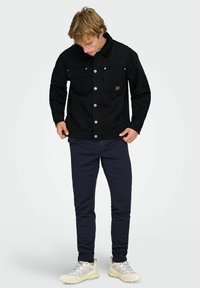 Črna denim jakna z ovratnikom iz korduroja, dvema sprednjima žepoma in srebrnimi gumbi; kombinirana z mornarsko modrimi hlačami in svetlimi supergami.