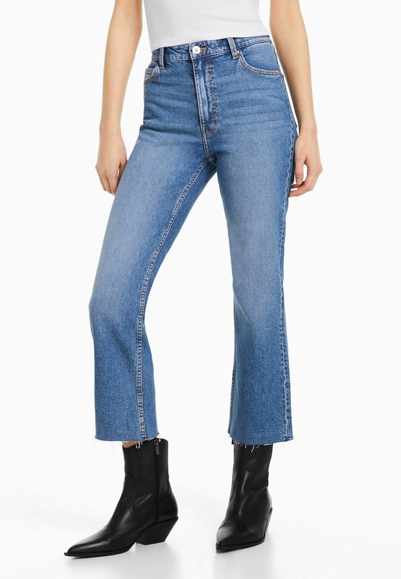 Femme portant un jean bleu court avec des ourlets bruts et des bottines noires, debout sur un fond blanc.