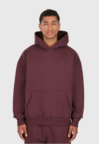 Giovane uomo che indossa un cappuccio color bordeaux scuro e pantaloni della tuta abbinati, in piedi contro uno sfondo chiaro e semplice, rivolto verso l'avanti.