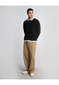 Camicia nera a maniche lunghe con colletto beige, abbinata a pantaloni beige a gamba larga e scarpe chiare. Dettaglio logo minimale sulla camicia.