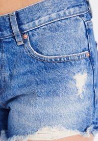 Detailní záběr na modré džíny šortky s prořezanými detaily a třepeným lemem na kyčli a horní části stehna osoby.