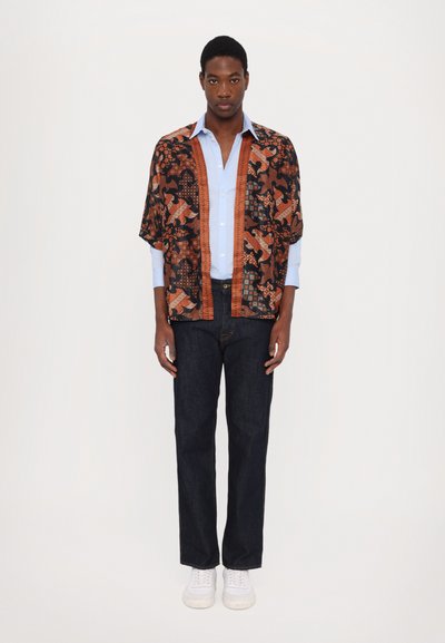 Schwarz- und orangefarbenes gemustertes Kimono über einem hellblauen Hemd, kombiniert mit dunklen Jeans und weißen Sneakern. Besitzt auffällige Grafiken und einen lockeren Schnitt.
