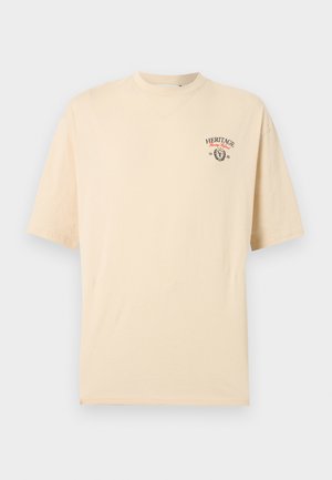 Beige T-shirt met korte mouwen, met Heritage-logo, rode letters en tekst "1982" op de bovenste linkerborst, weergegeven op een effen achtergrond.