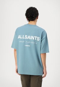 T-shirt oversize bleu clair en coton, avec un grand texte blanc "ALLSAINTS" et un texte supplémentaire en japonais ainsi que "LONDON" au dos.